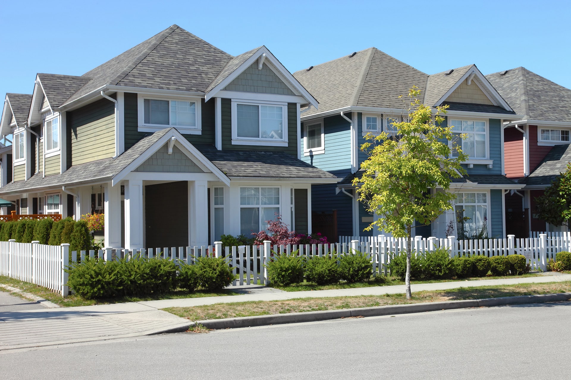 Row of suburban houses with pitched roofs and white picket fences, surrounded by greenery on a clear day.