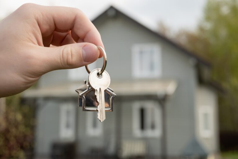 Man hand holds house-shaped key in front of newly purchased home.